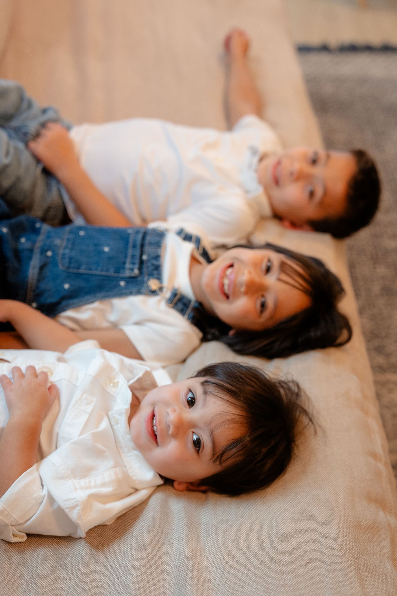 Three young children lying together on a beige cushion, smiling at the camera in a cozy indoor setting.