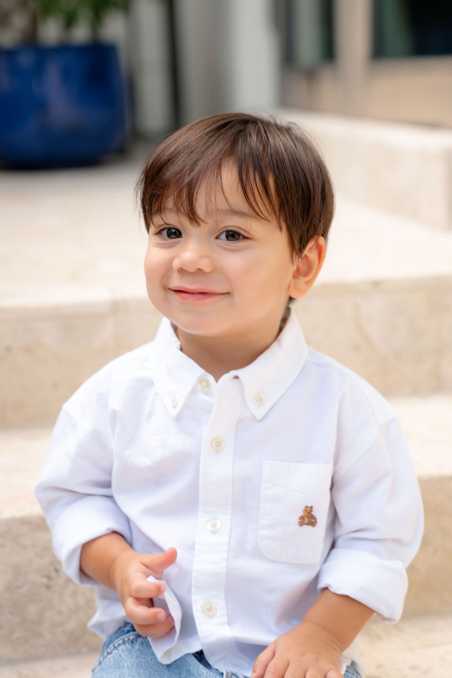 Smiling toddler boy in white button-up shirt with bear logo sitting on steps outdoors.