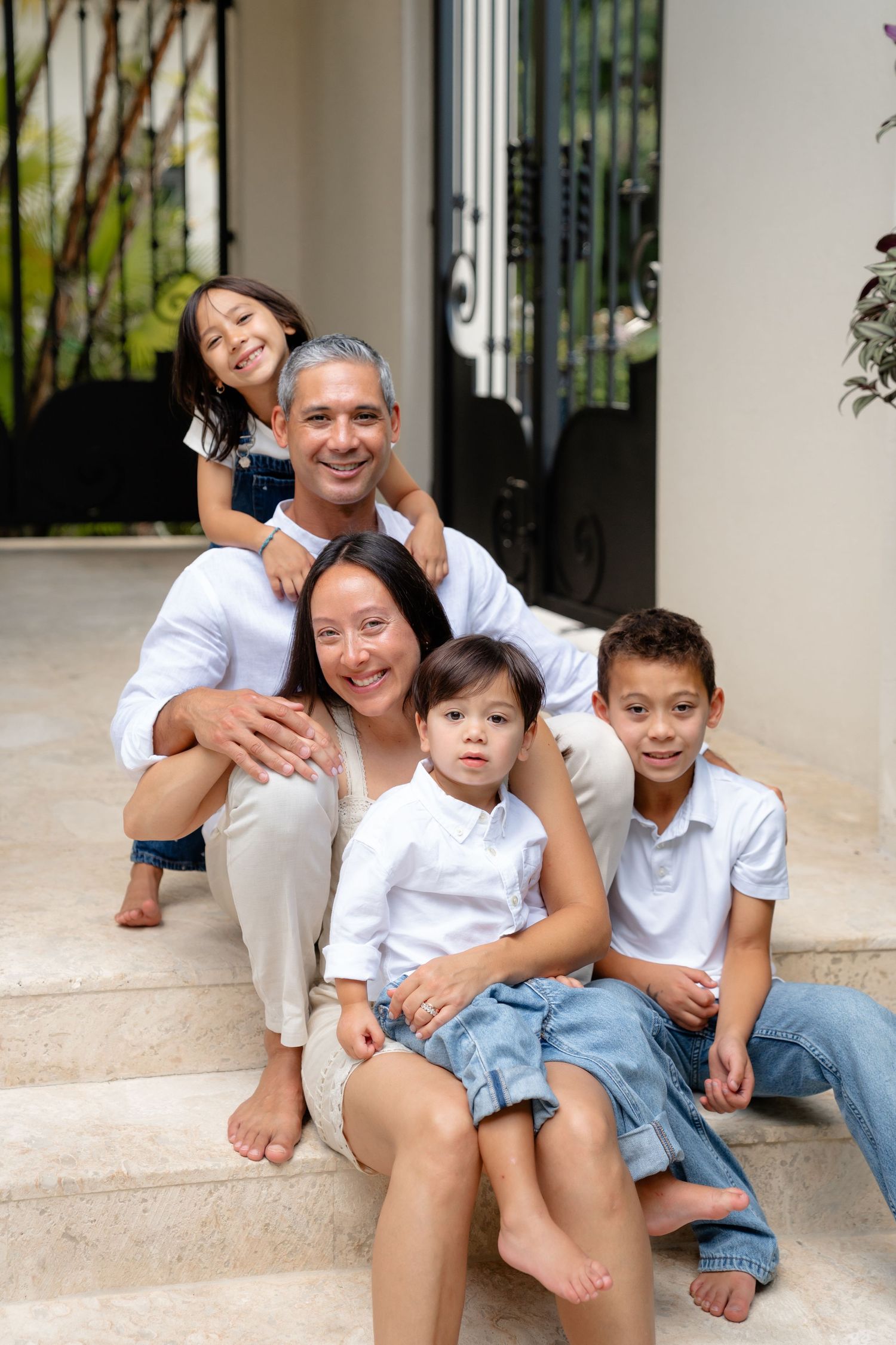 Happy multiracial family of five smiling together on home entrance steps, parents with three young children in white outfits.