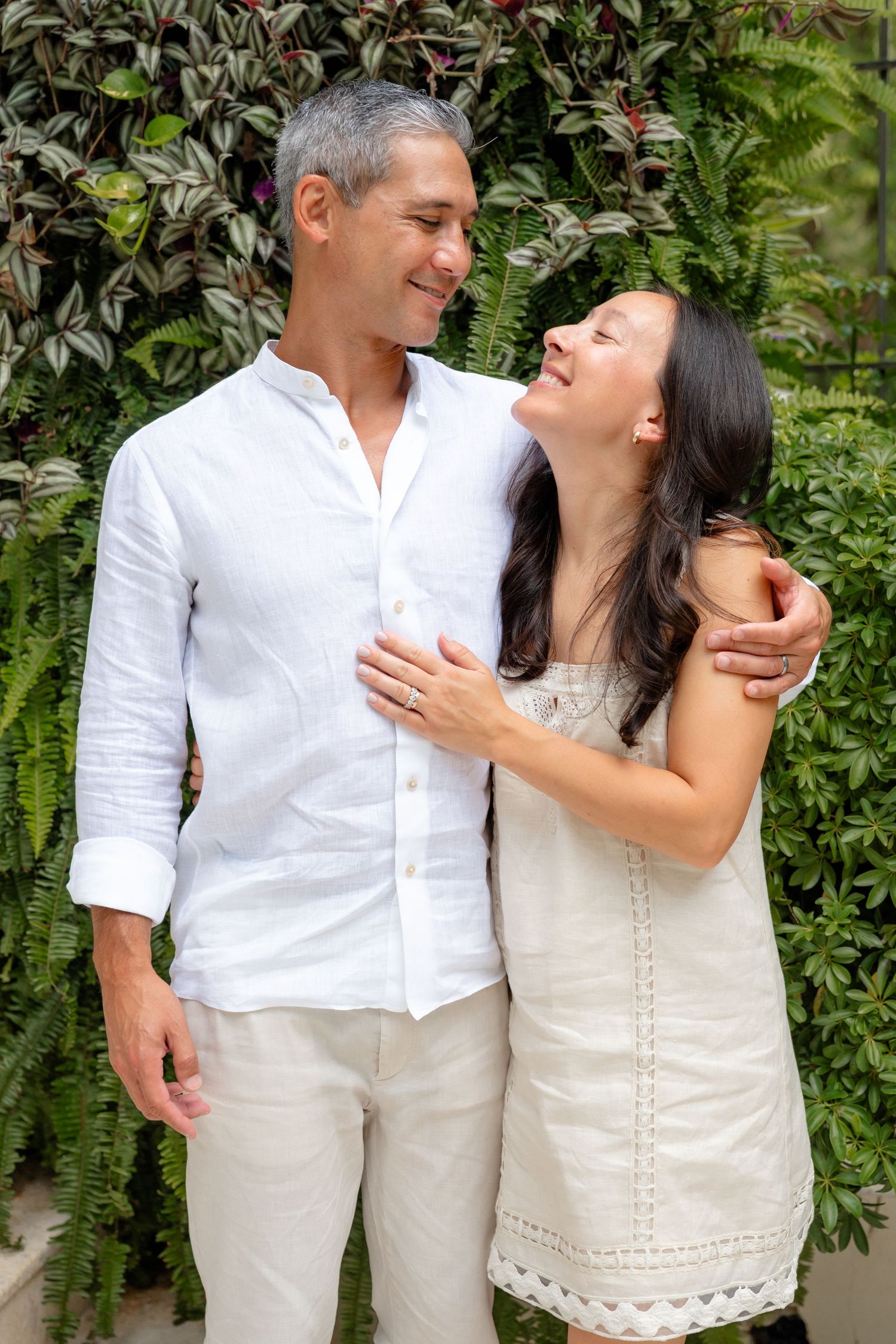 A smiling couple in white outfits embracing outdoors in front of lush green tropical foliage.