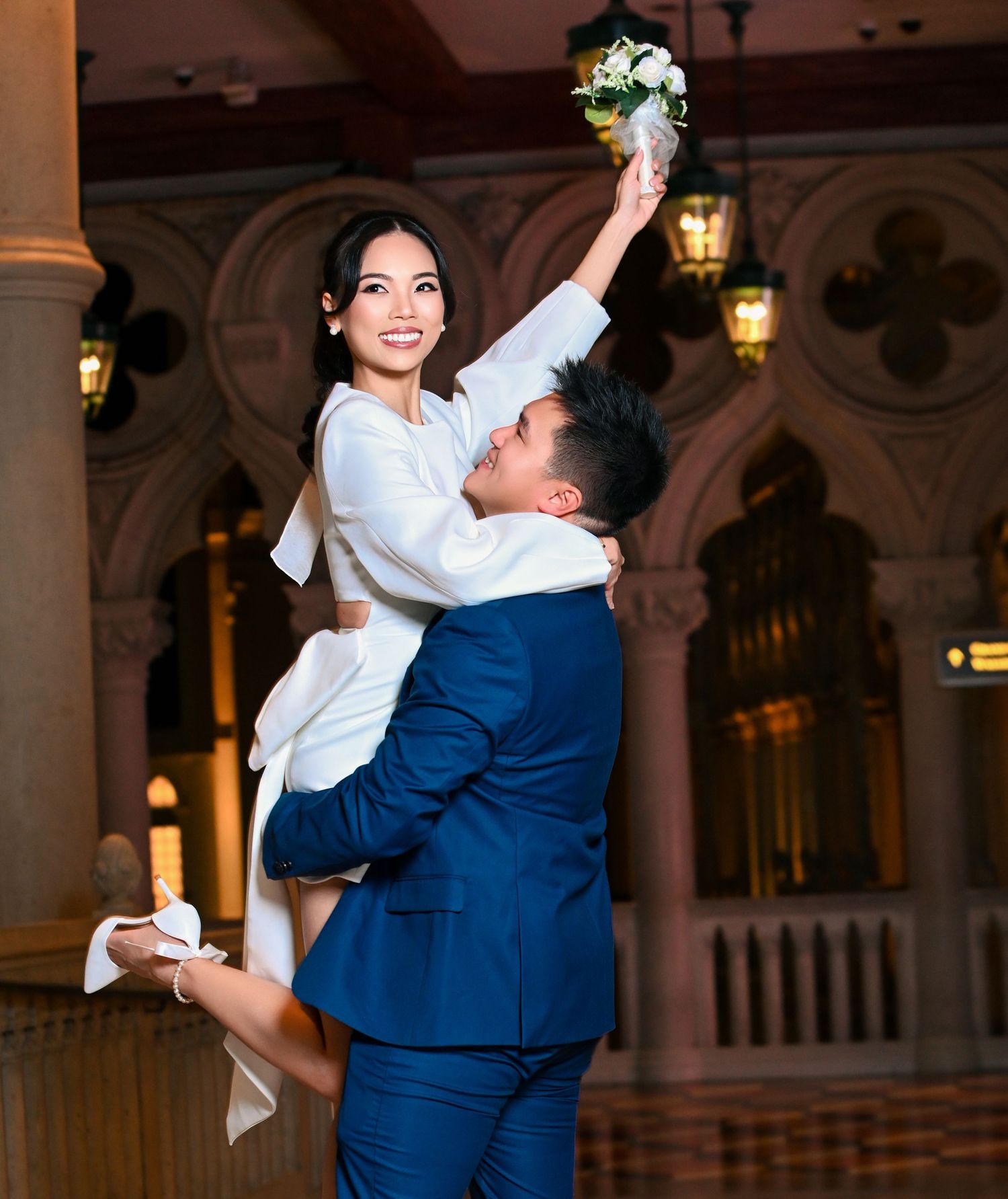 Groom in blue suit lifts bride in white dress holding bouquet aloft in elegant wedding venue.