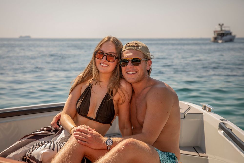 A happy couple sitting together on the edge of a luxury boat, smiling and looking directly at the camera with the sparkling sea in the backg