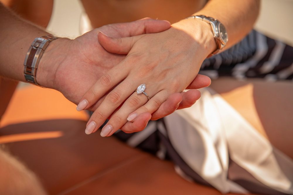 Close-up of a woman's hand resting on top of a man's hand, showcasing a brilliant oval-cut diamond engagement ring.
