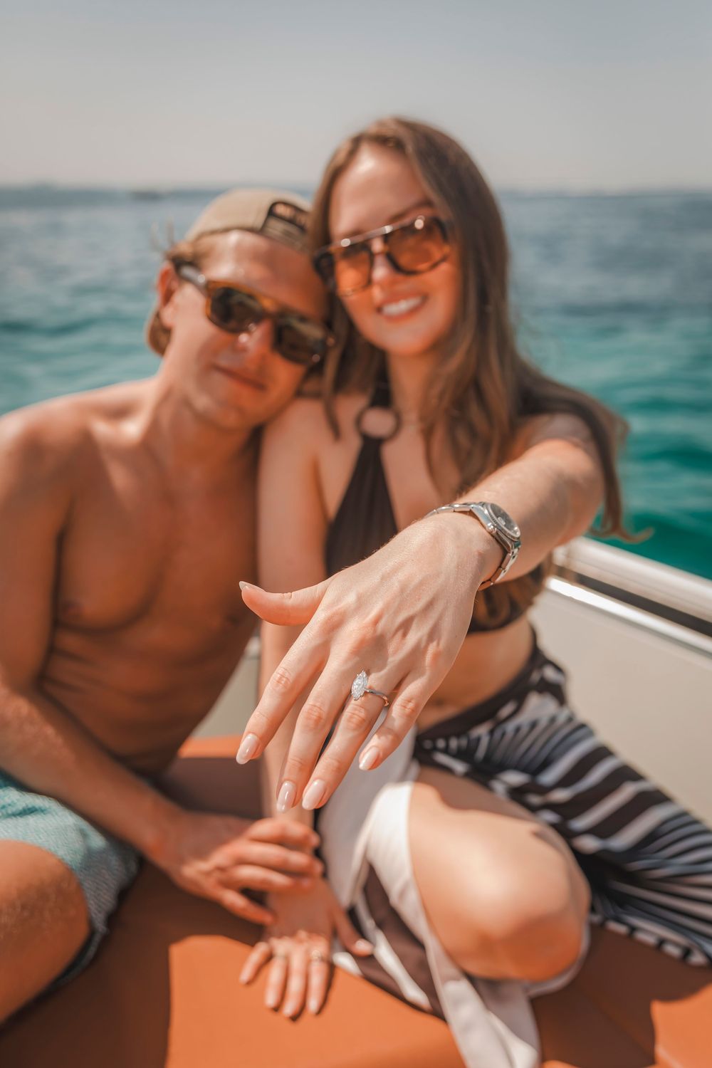 A couple sitting together on a boat, with the woman holds her hand out toward the camera to display her new engagement ring.