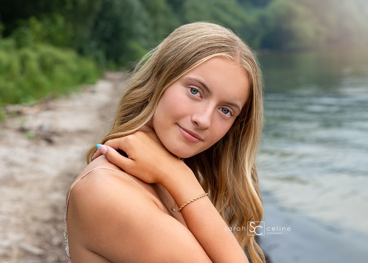 Teen girl with long blonde hair poses by a river, smiling softly in a natural outdoor setting.