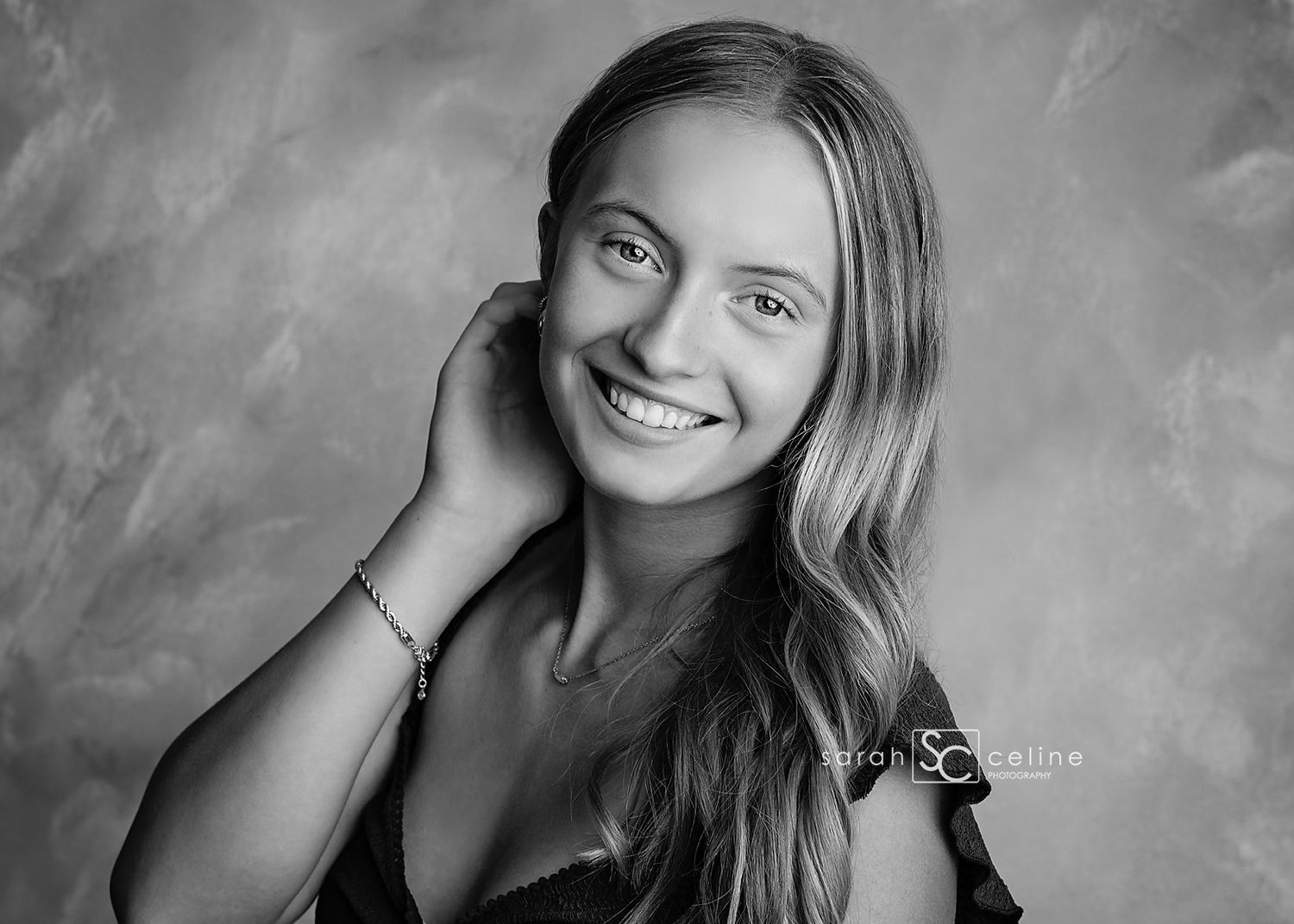 Black and white senior portrait of a smiling young woman with long wavy hair, posing gracefully against a textured backdrop.