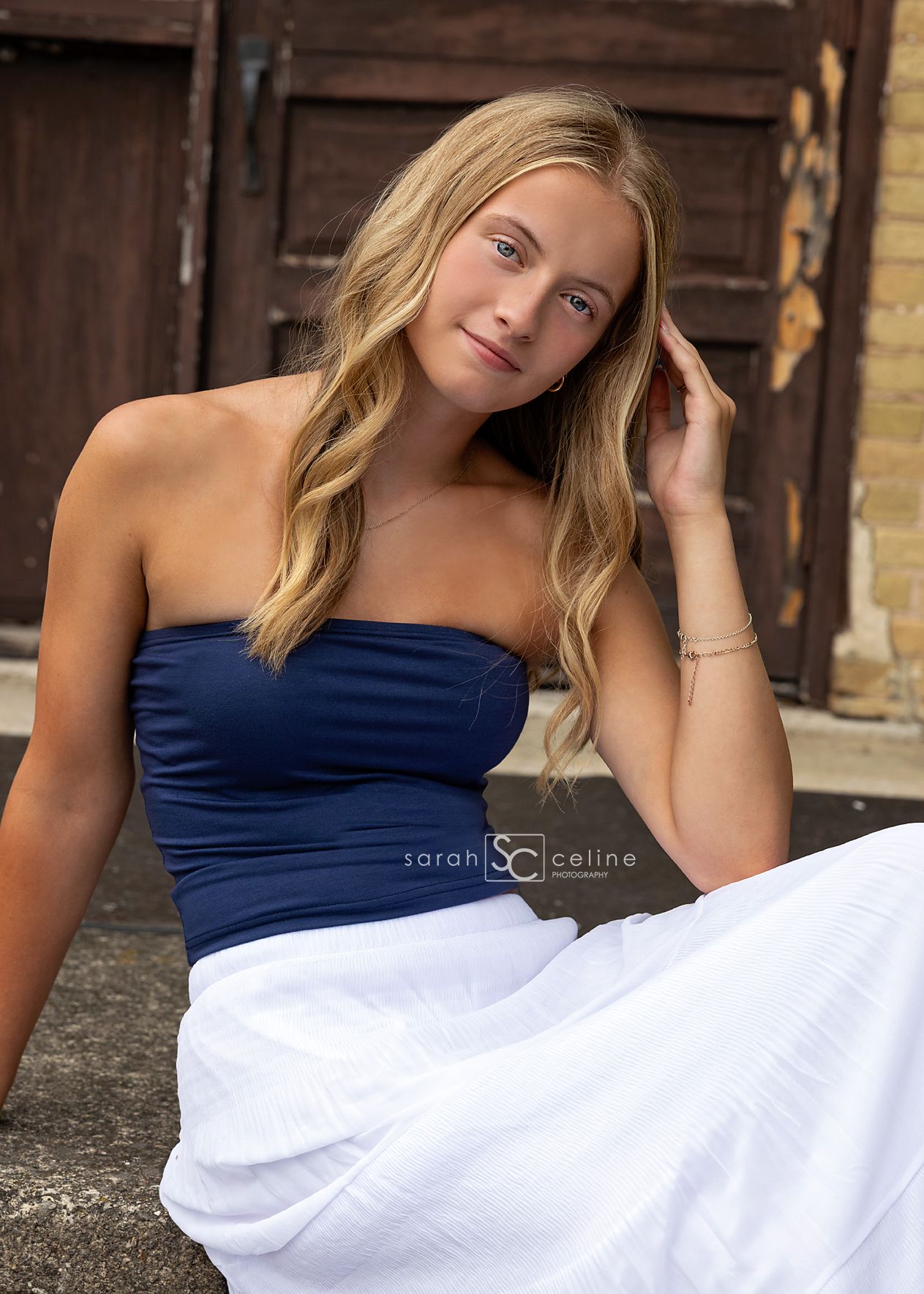 Young woman in navy strapless top and white skirt poses outdoors near rustic wooden doors for a senior portrait.