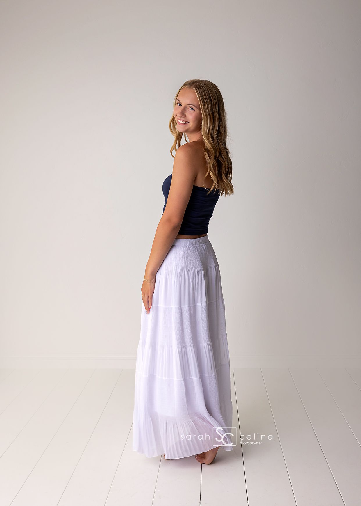 Teen girl in navy crop top and white maxi skirt smiling over shoulder in studio portrait.