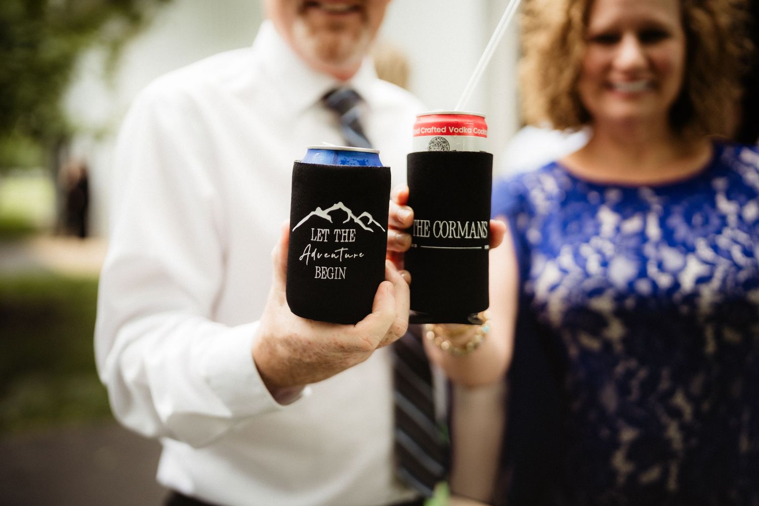 Couple holding custom black wedding koozies reading Let the Adventure Begin and The Corman at outdoor ceremony.