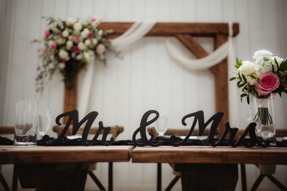 Elegant wedding sweetheart table with black Mr. & Mrs. signs, floral arrangements, and rustic wooden arch backdrop.
