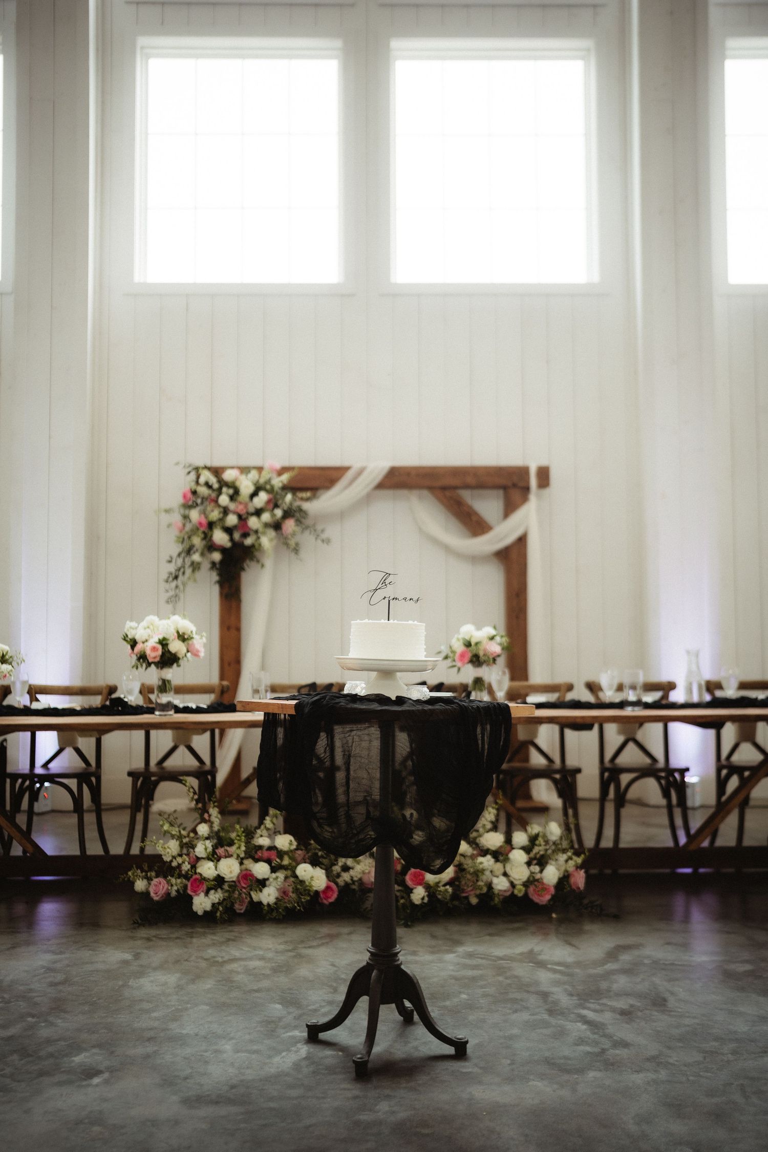 Elegant wedding reception with floral arch, white cake on black pedestal table, and long wooden head table in bright barn venue.