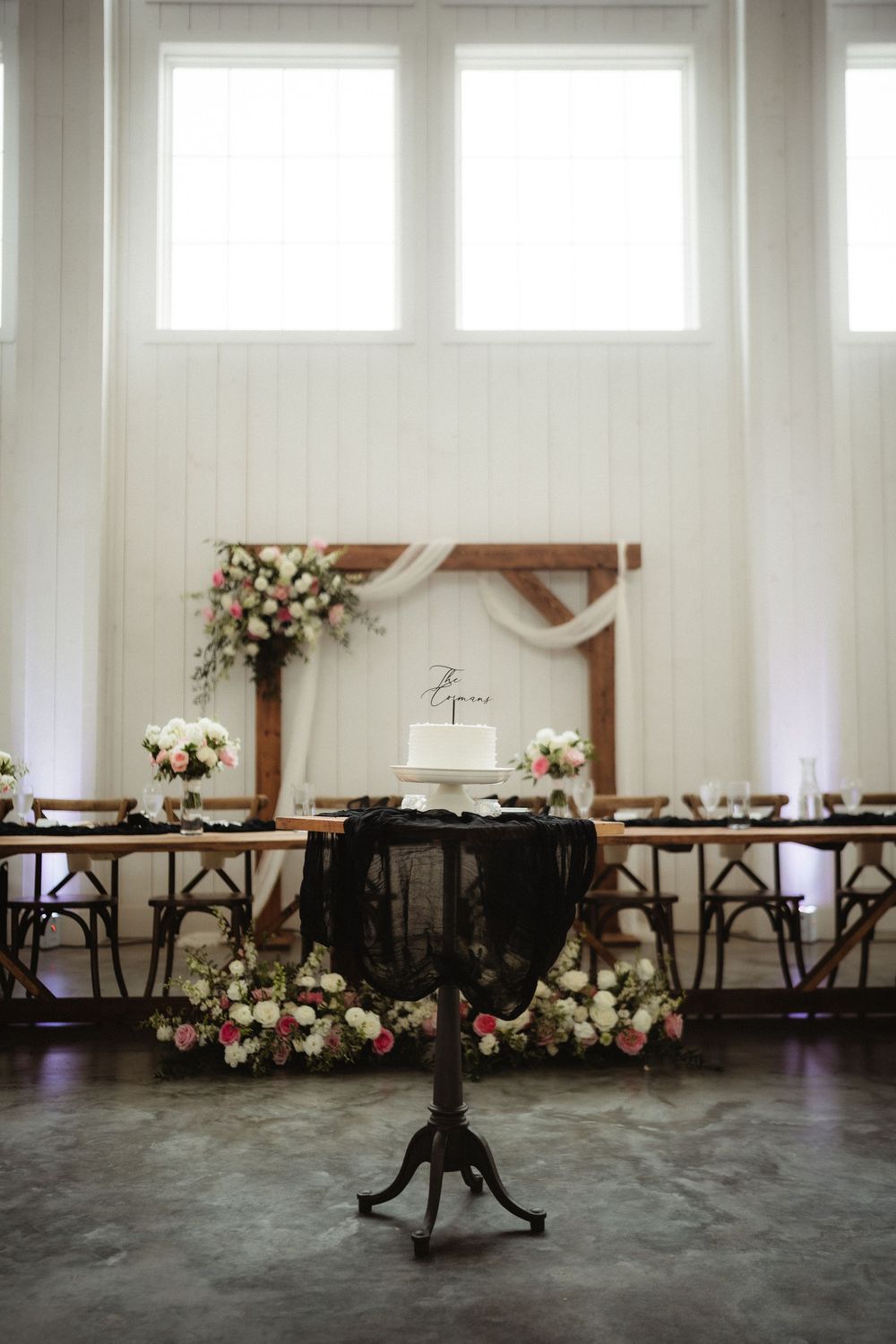 Elegant wedding reception with floral arch, white cake on black pedestal table, and long wooden head table in bright barn venue.