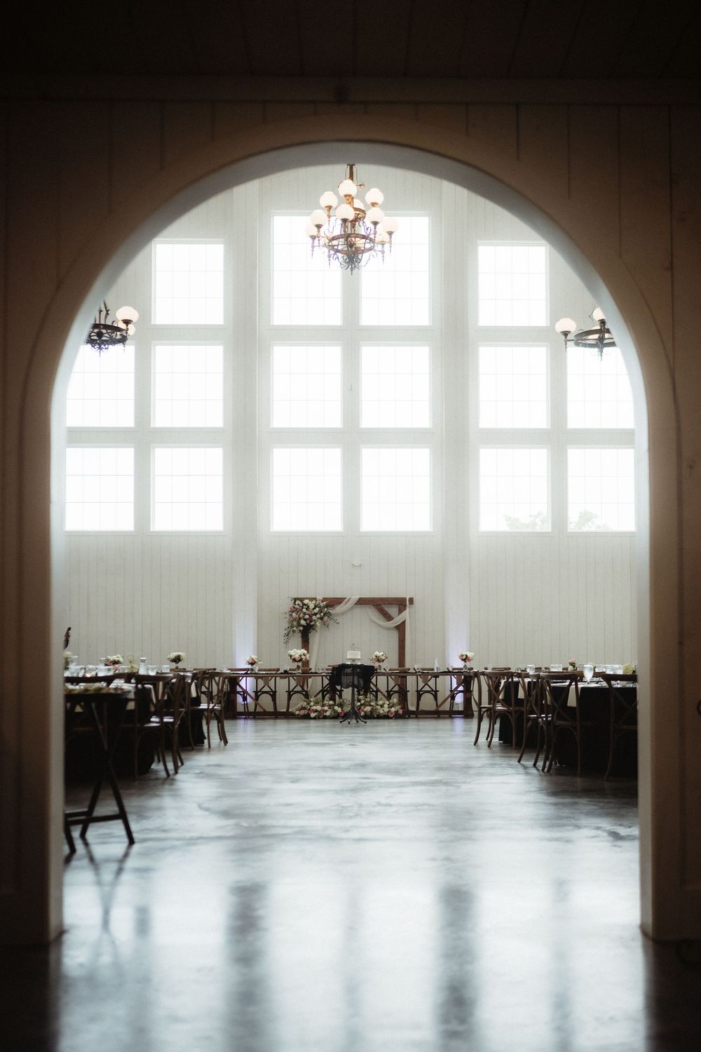 Elegant wedding reception hall viewed through arched doorway with chandelier and large windows.