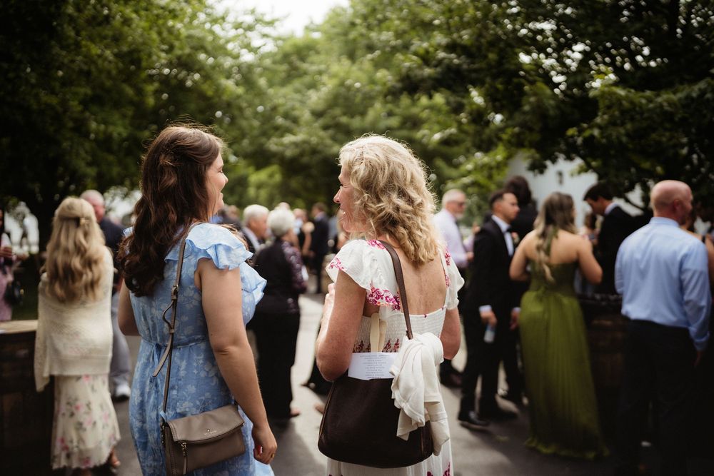 Two women in floral dresses chat at an outdoor garden party with guests mingling in the background.