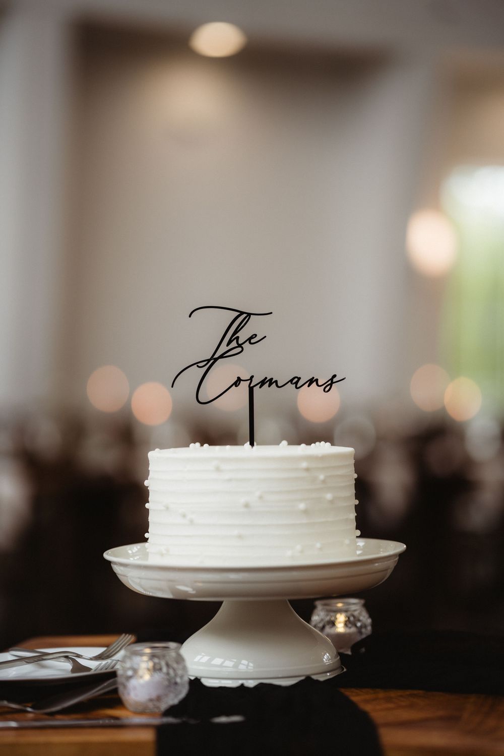 Simple white wedding cake with pearl details and The Coimans custom black script cake topper on pedestal.