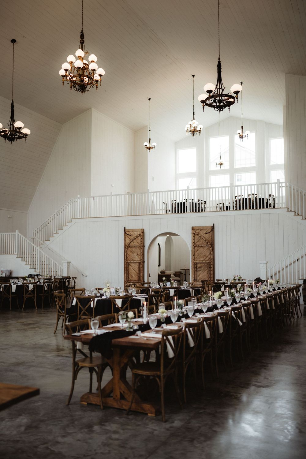Spacious wedding venue with long farm tables, chandeliers, and grand staircase in white hall.
