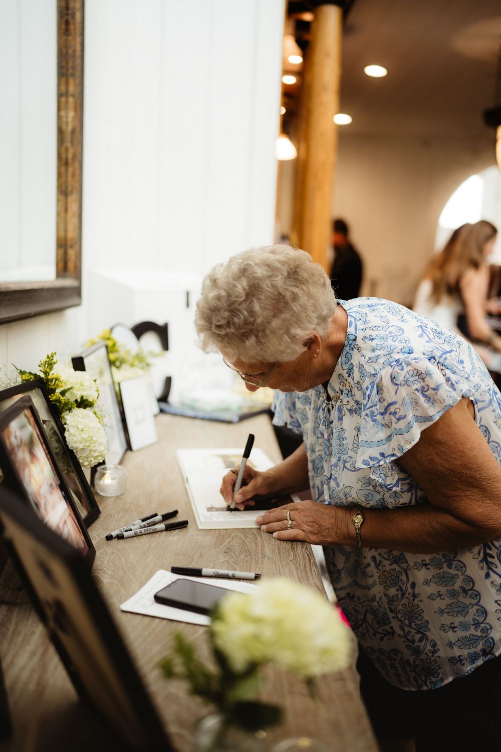 Elderly woman in floral blouse signing guest book at wooden table decorated with white hydrangeas at wedding reception.