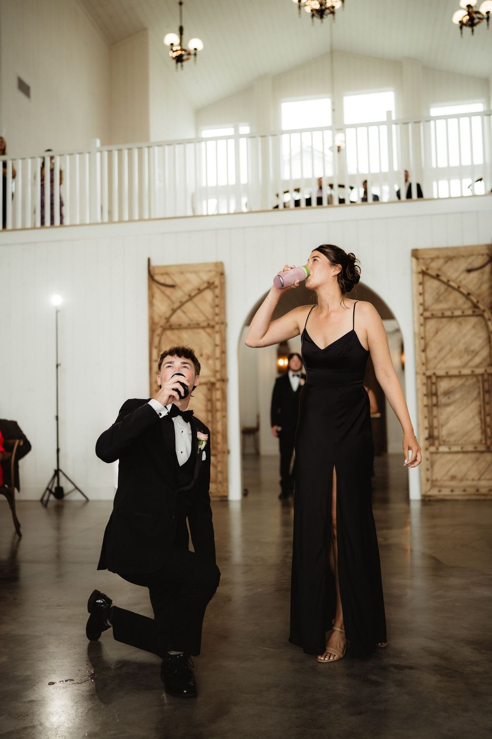 Wedding guests in black formal attire drinking and celebrating on dance floor of white barn venue.