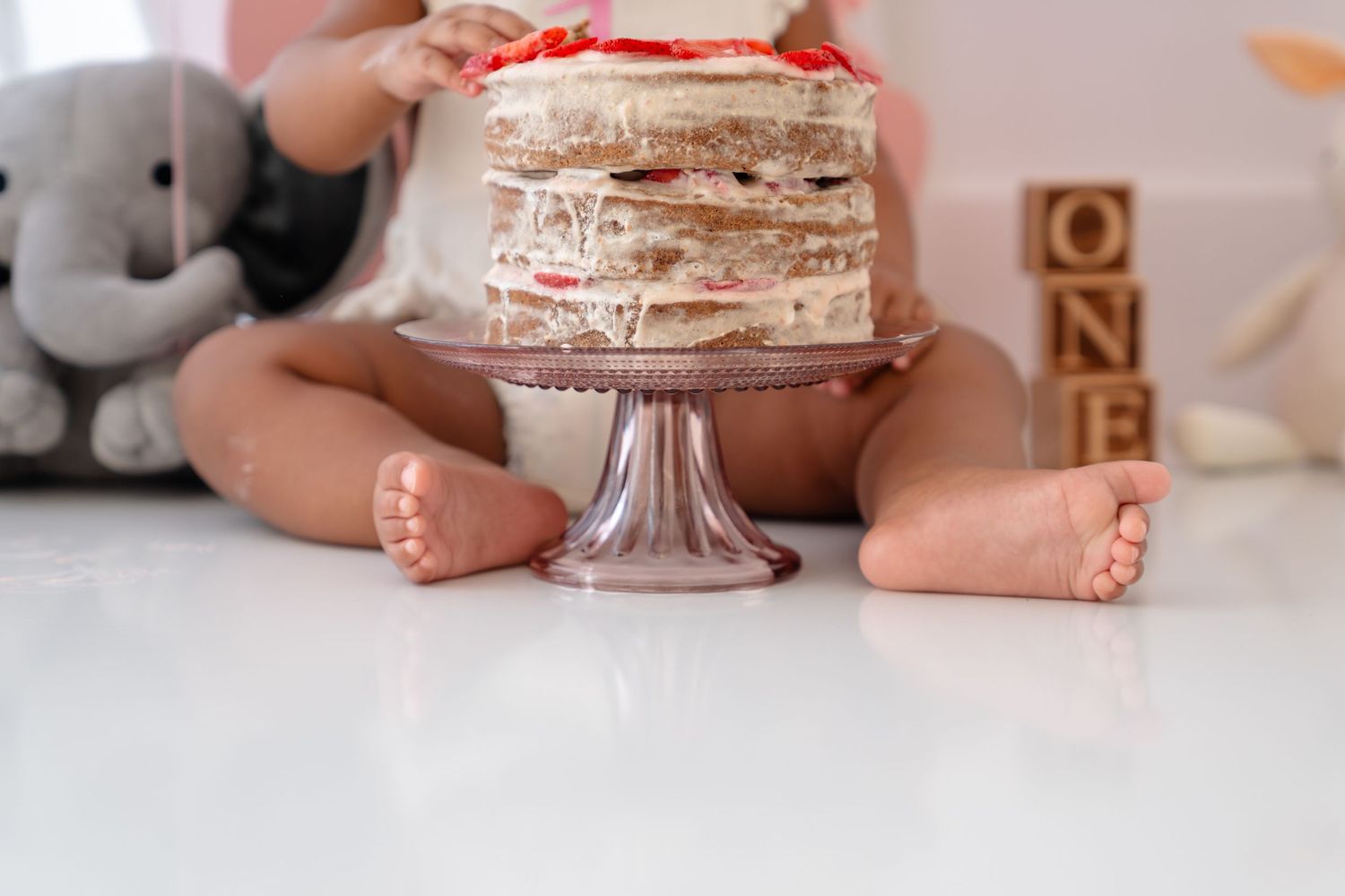 Baby's first birthday cake smash with layered naked cake on glass stand, wooden ONE blocks, and stuffed elephant toy.