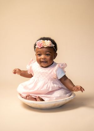 Smiling baby girl in pink smocked dress and floral headband sitting in a bowl, studio portrait on beige background.