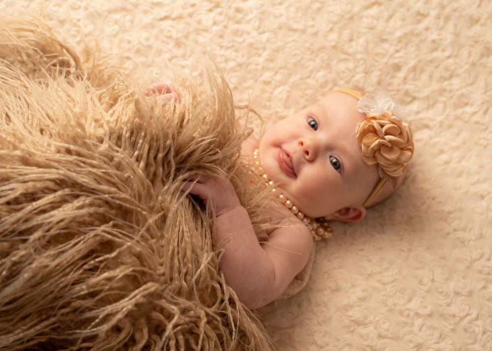 Adorable baby girl lying on a fluffy cream rug wearing a pearl necklace and floral headband.