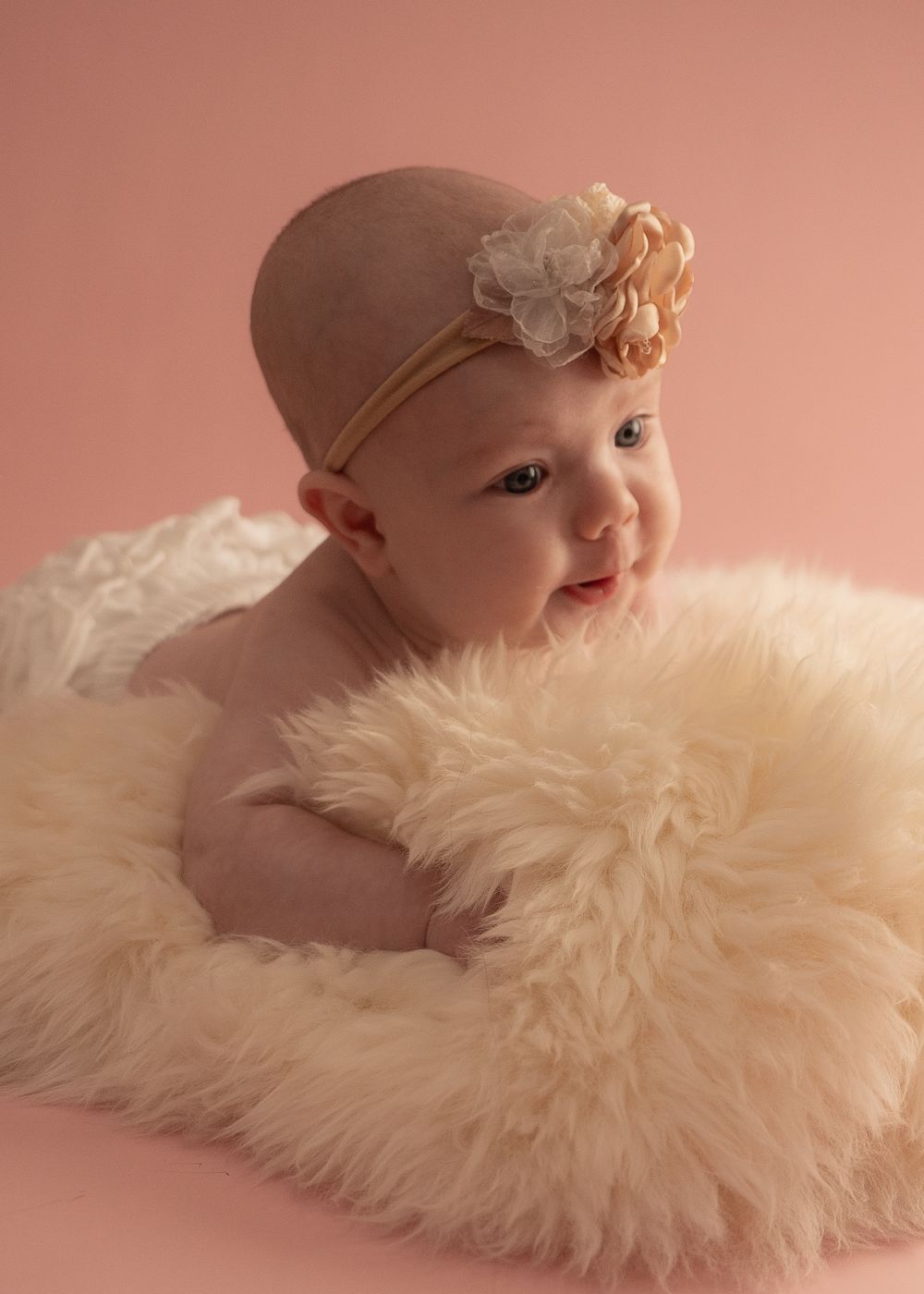 Adorable baby girl in a fluffy cream fur nest wearing a floral headband, posed on a pink background.