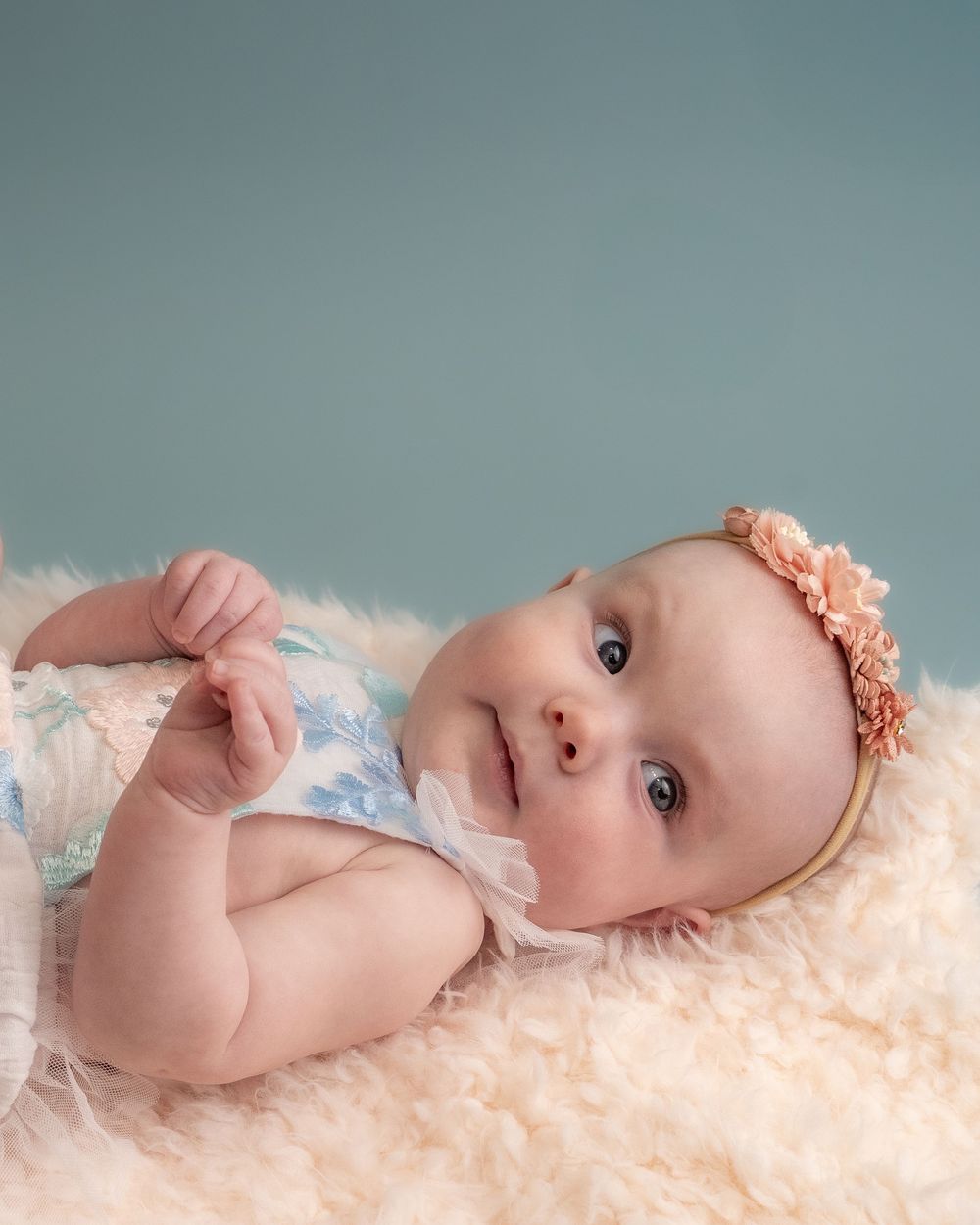 Adorable baby girl in floral romper and pink flower headband lying on fluffy cream rug with teal background.