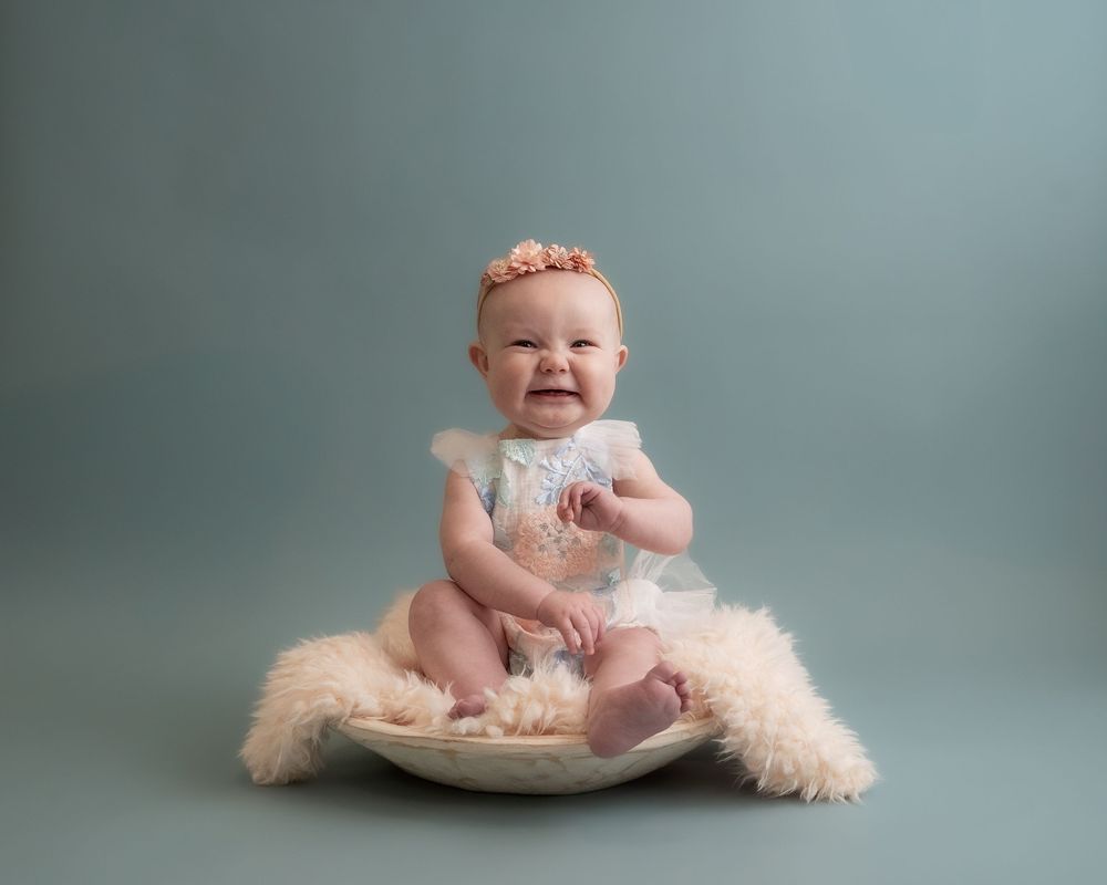 Smiling baby girl in white lace dress and floral headband sitting in a bowl on fluffy rug, studio portrait.