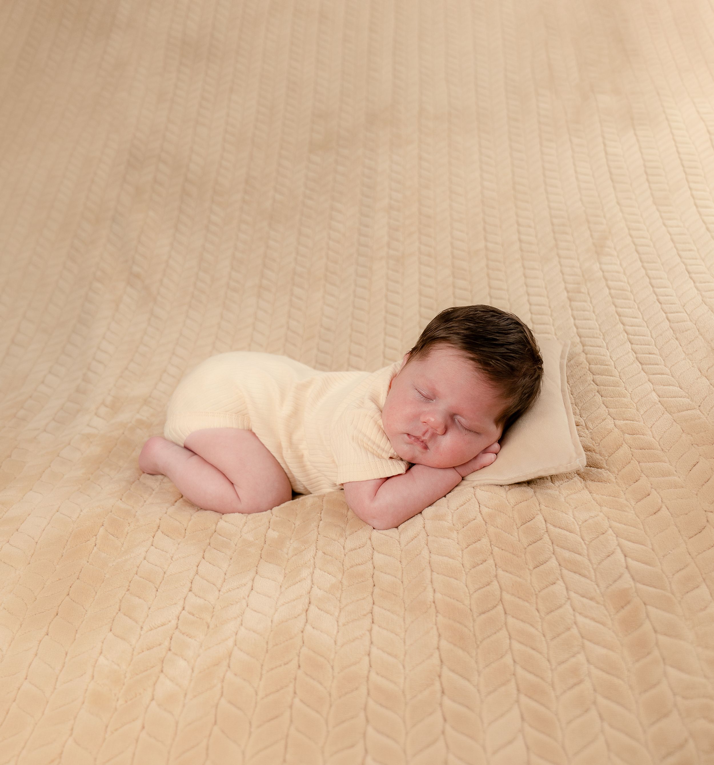 Newborn baby sleeping peacefully on a beige textured blanket wearing a soft yellow onesie with dark hair.