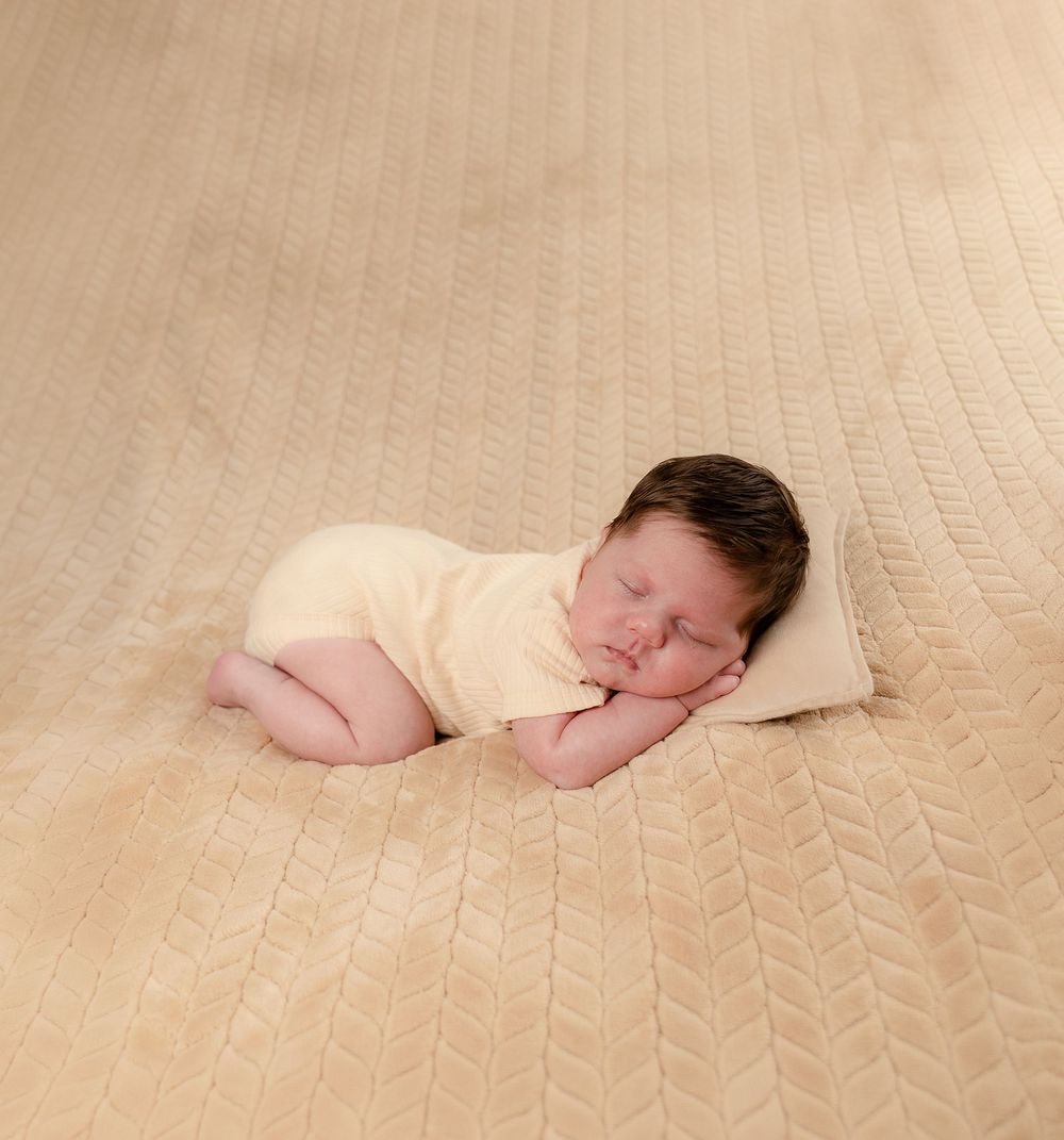 Newborn baby sleeping peacefully on a beige textured blanket wearing a soft yellow onesie with dark hair.