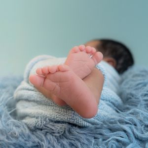 Close-up of a sleeping newborn baby's tiny feet wrapped in a soft blue knit blanket on a fluffy blue surface.