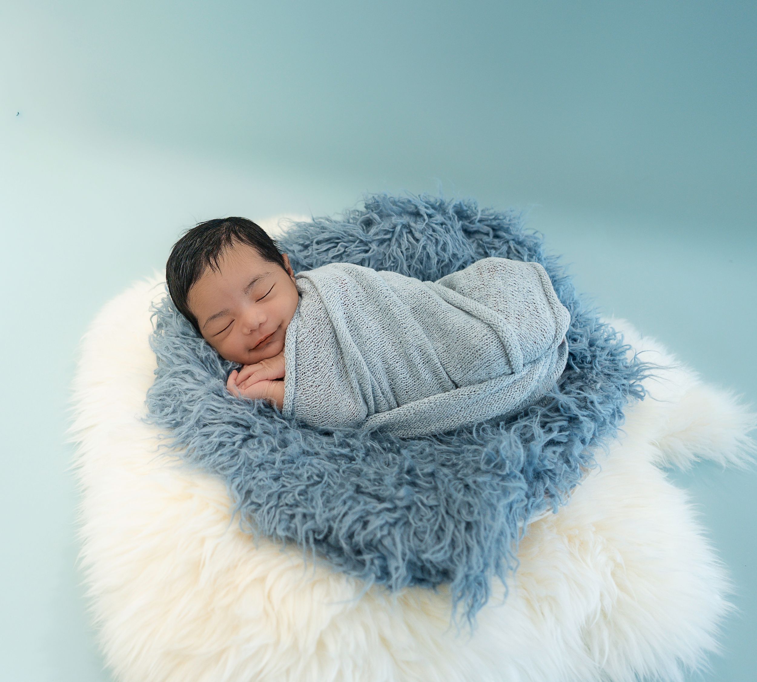 Peaceful newborn baby sleeping in a blue fuzzy nest wrap on a white fluffy blanket with a soft blue background.