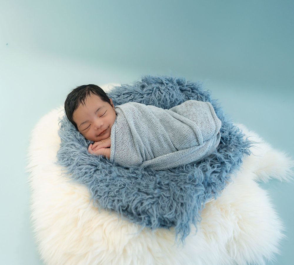 Peaceful newborn baby sleeping in a blue fuzzy nest wrap on a white fluffy blanket with a soft blue background.