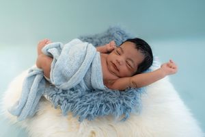 Smiling newborn baby wrapped in a soft blue knit blanket, resting on a fluffy white fur rug in a studio.