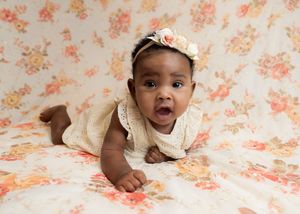Adorable dark-skinned baby girl in cream lace romper and floral headband doing tummy time on floral backdrop.