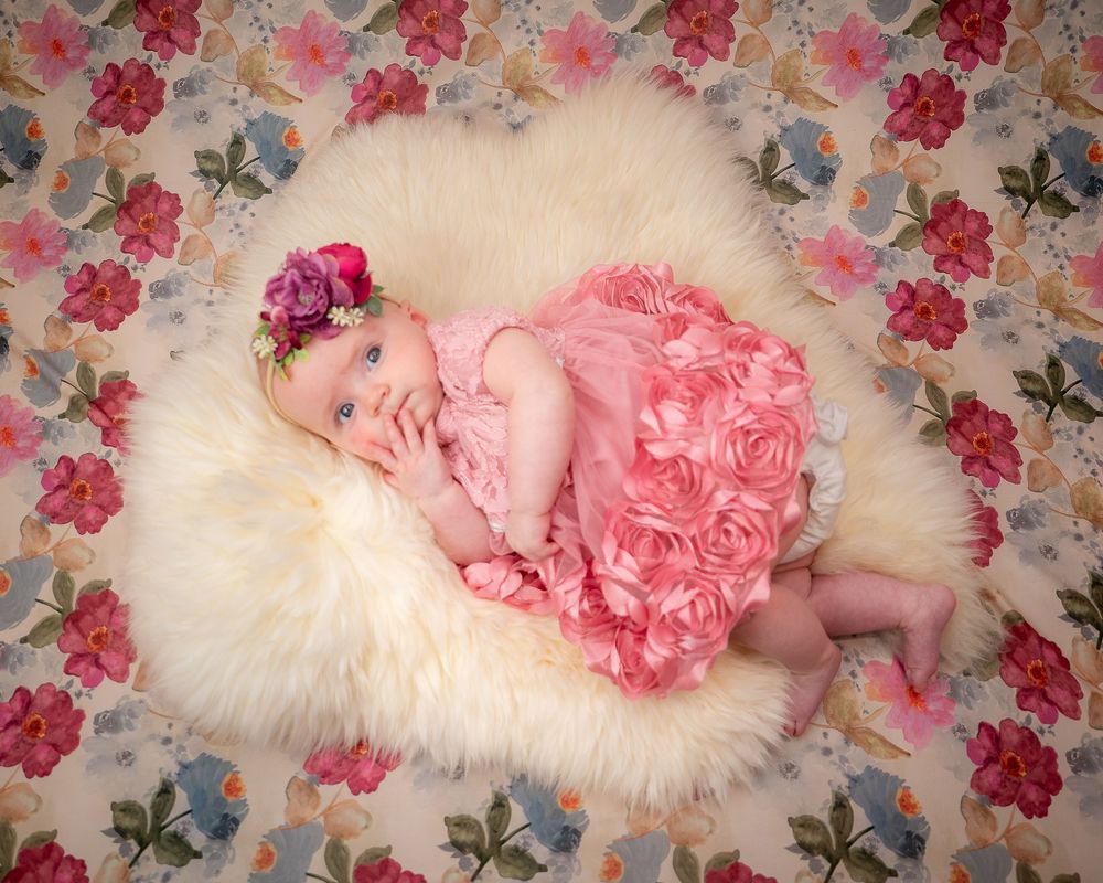 A smiling baby girl in a pink rosette dress and floral headband lies on a fluffy white rug with a floral backdrop.