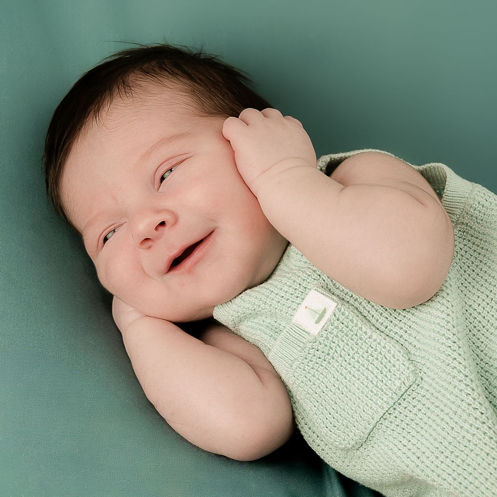 Smiling newborn baby in a mint green knit romper lying on a teal background.
