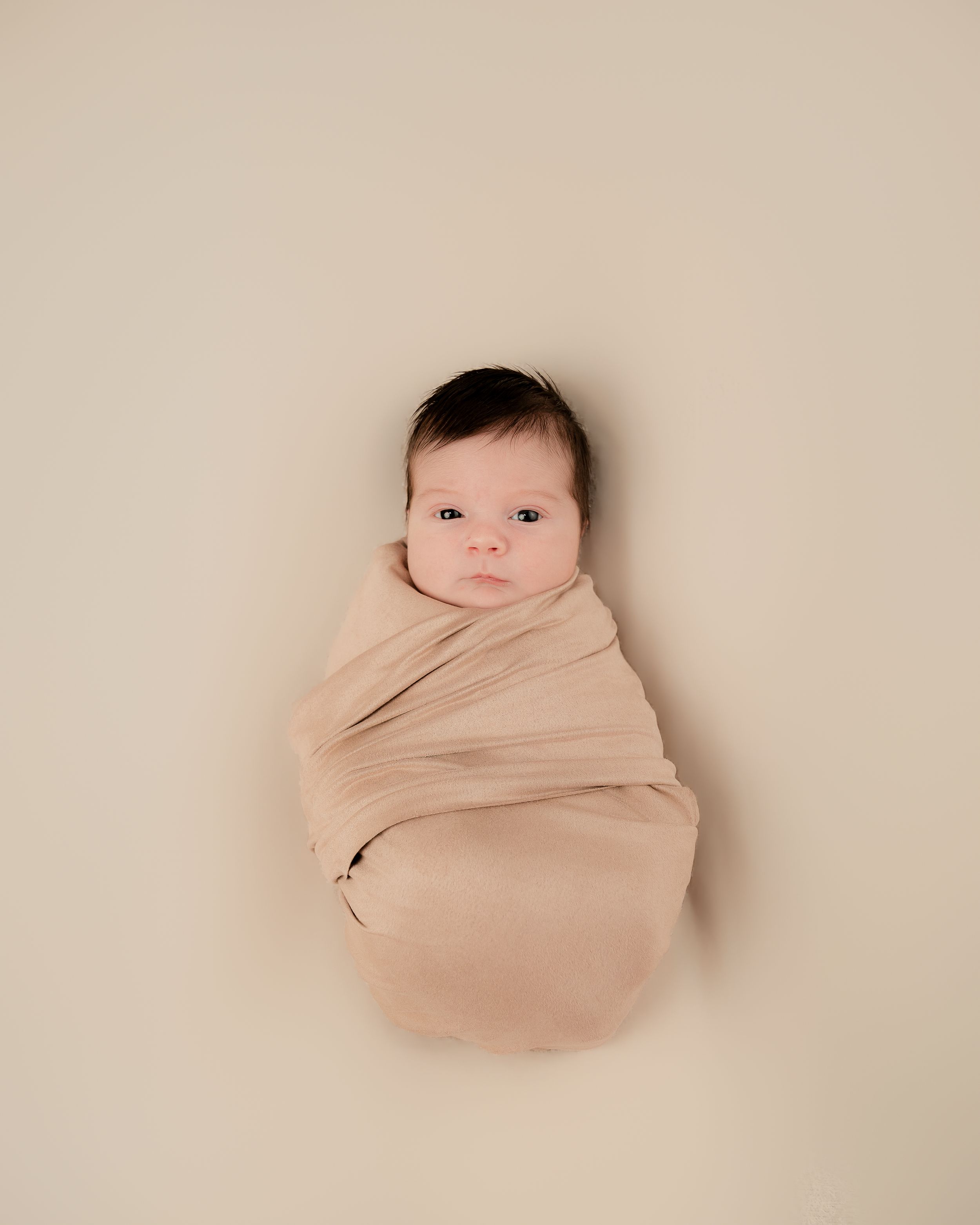 Newborn baby swaddled in a soft beige wrap, lying on a neutral background in a professional studio portrait.
