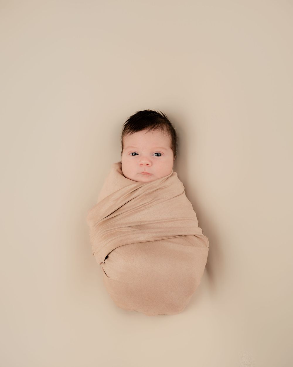 Newborn baby swaddled in a soft beige wrap, lying on a neutral background in a professional studio portrait.