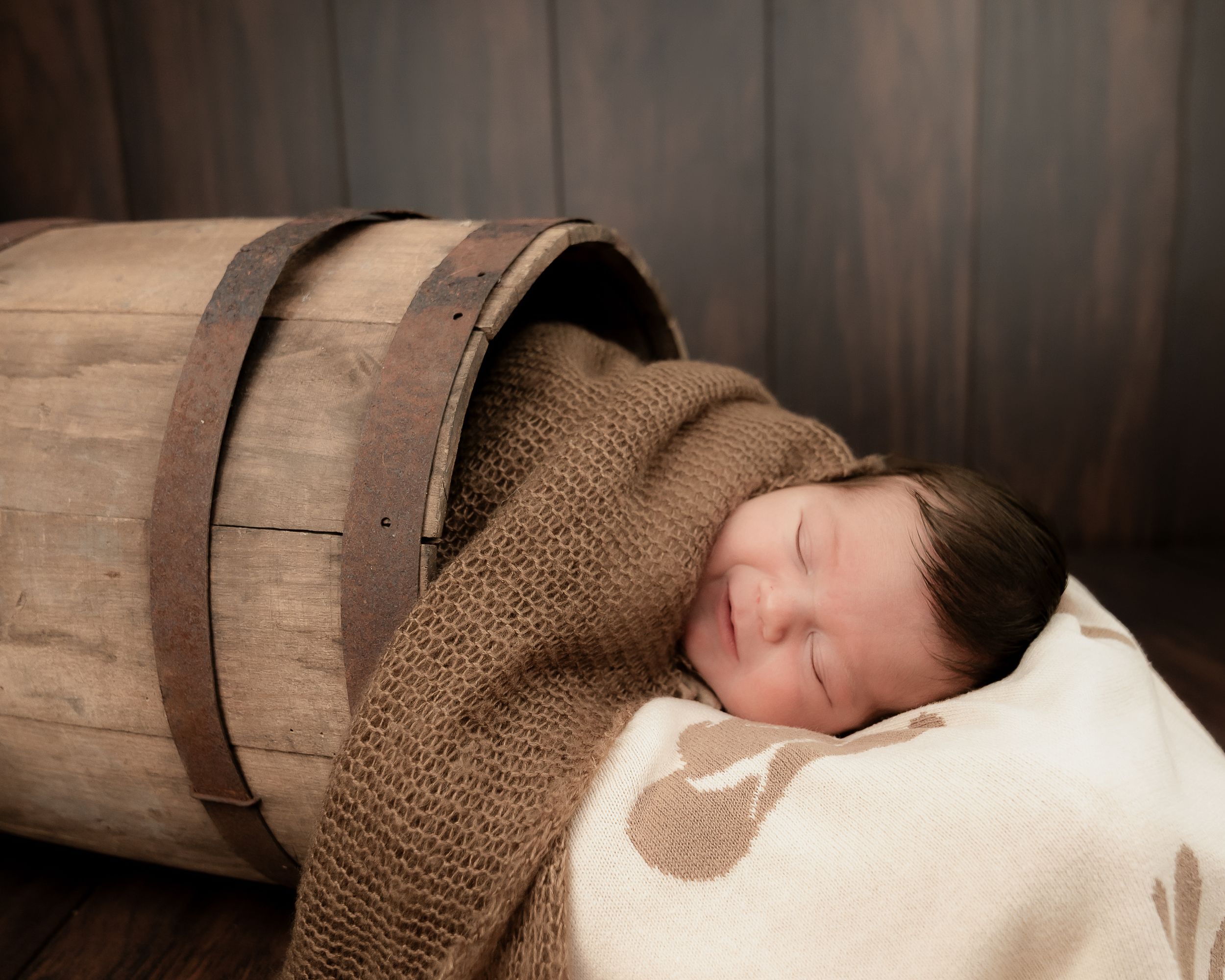 Sleeping newborn baby wrapped in brown knit blanket nestled inside rustic wooden barrel prop.