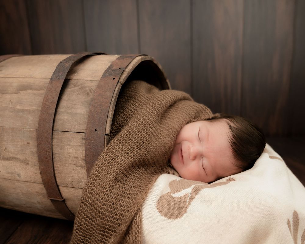 Sleeping newborn baby wrapped in brown knit blanket nestled inside rustic wooden barrel prop.