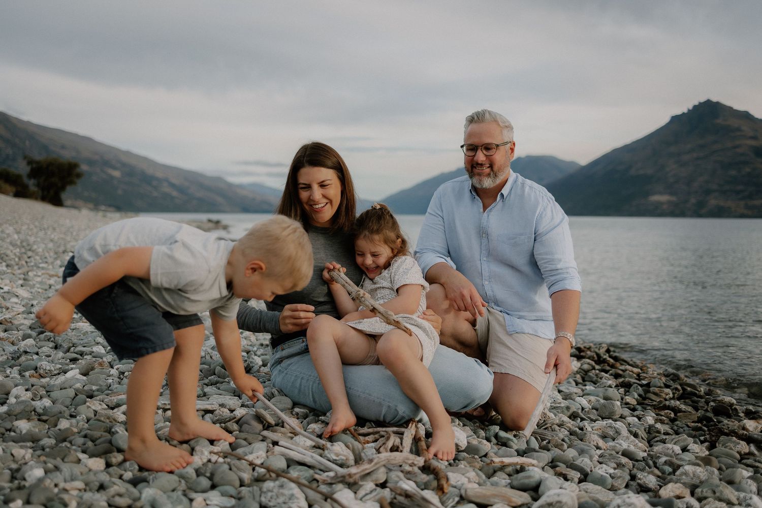 Family of four enjoying a rocky beach by a mountain lake, laughing together on a cloudy day.