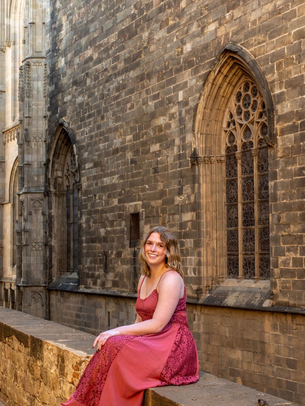 A woman in a red dress sits gracefully beside a Gothic stone cathedral in Barcelona's historic quarter.
