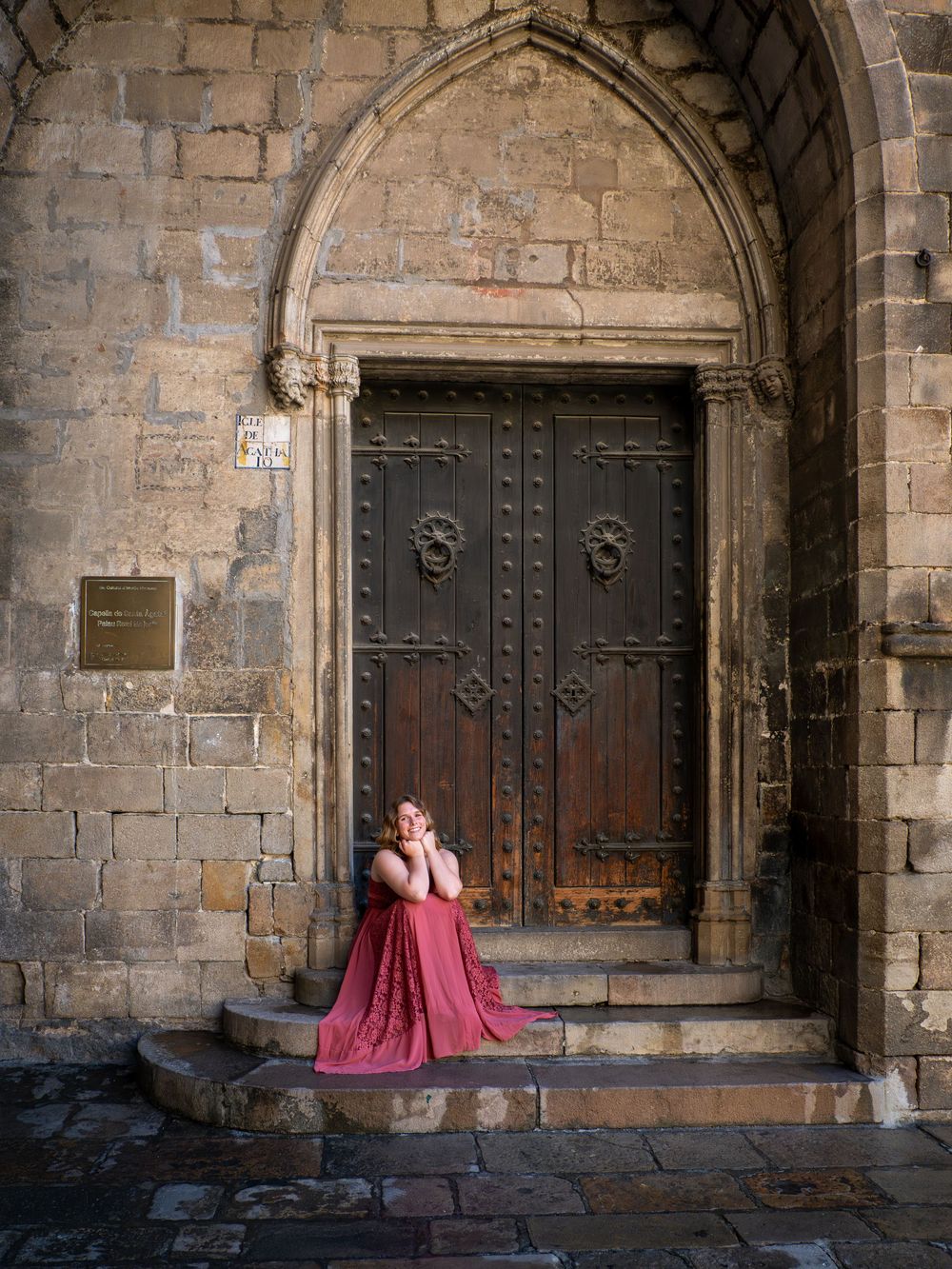 Woman in red gown sitting on stone steps before ornate medieval wooden door in historic Barcelona.