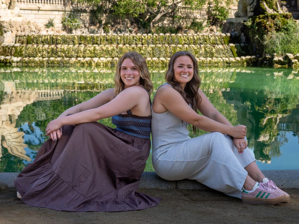Two young women sitting back-to-back by a green reflective pool in a lush garden setting.
