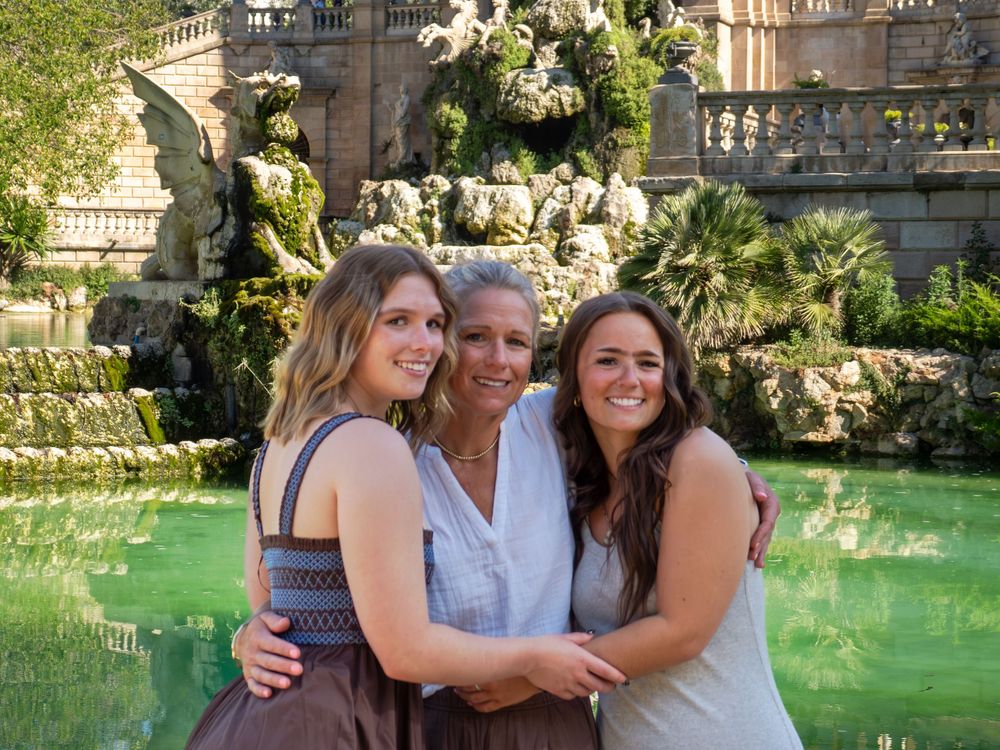 Three women smiling and embracing in front of an ornate fountain with green water and lush greenery.