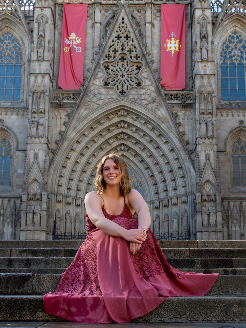 Young woman in elegant burgundy gown poses on cathedral steps with Gothic architecture and red banners behind her.