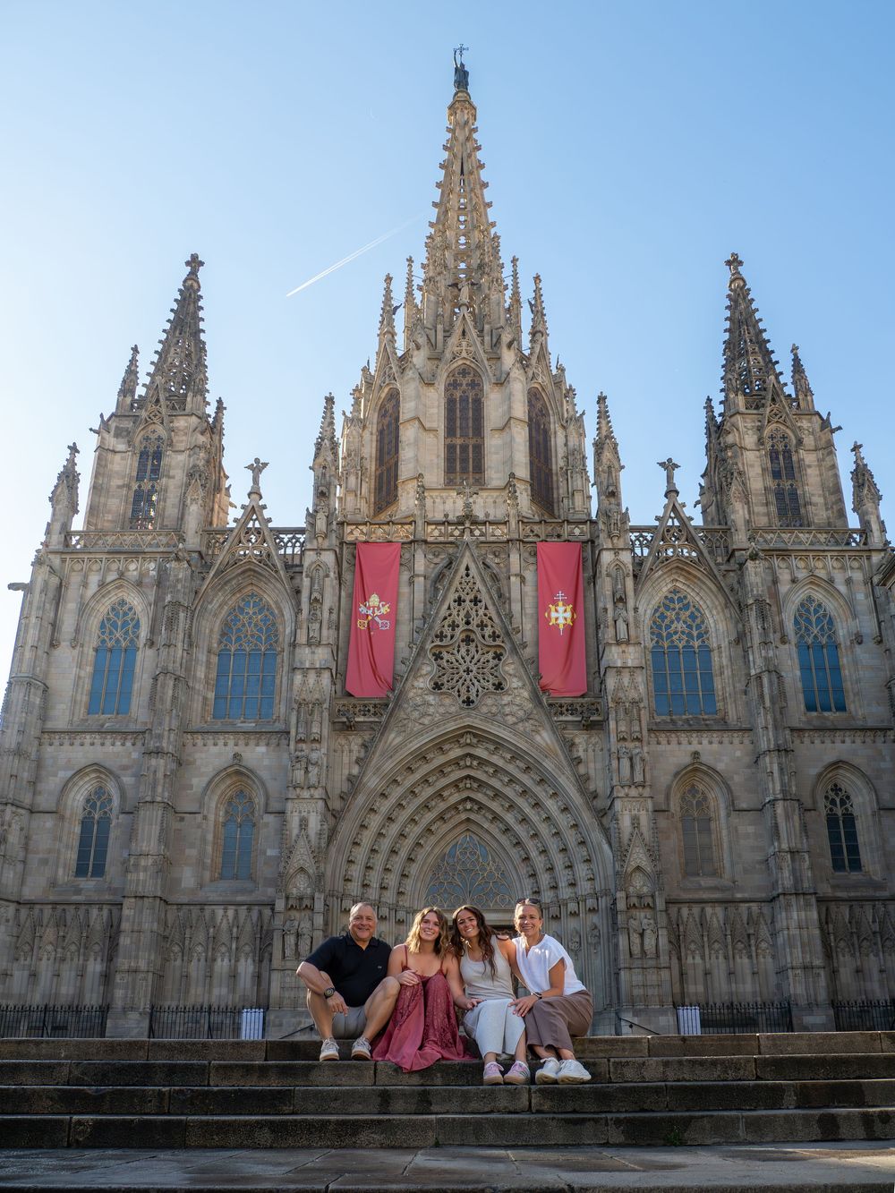 Four people sitting on steps before Barcelona Cathedral's Gothic facade with red banners, under clear blue sky.