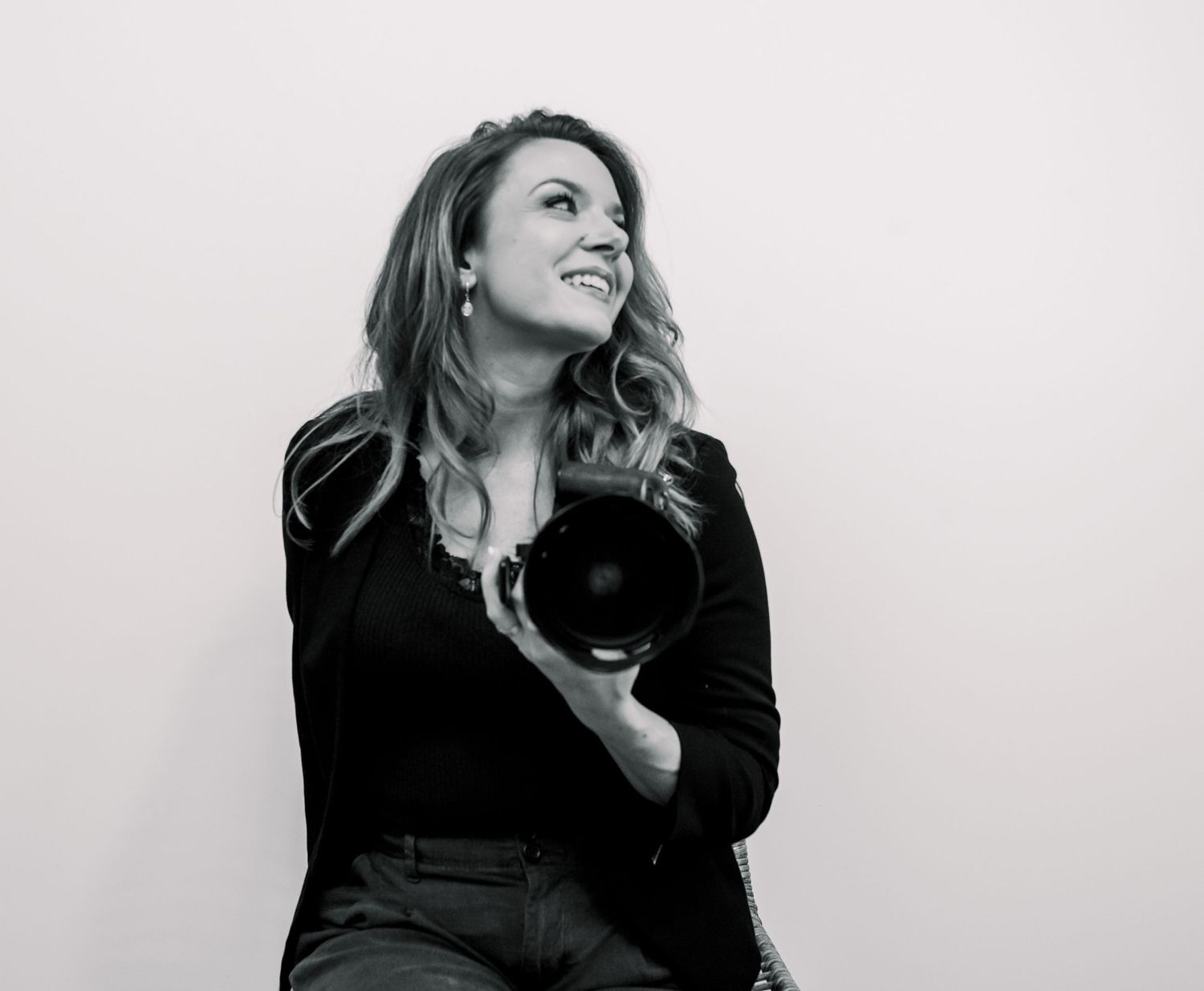 Black and white photo of a smiling woman holding a camera, wearing a black blazer against a light background.