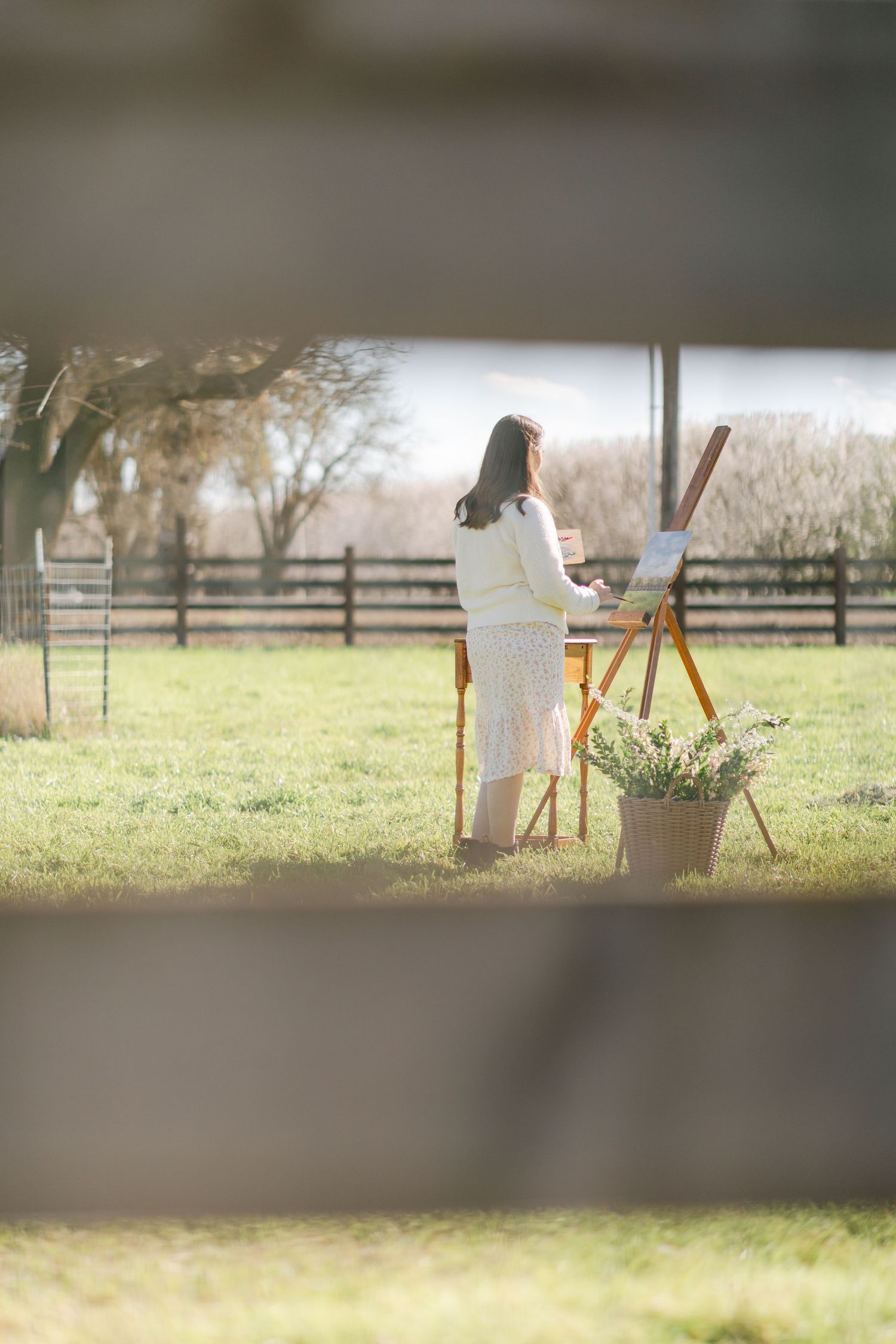 Woman painting on an easel outdoors in a sunny farm field, viewed through a rustic wooden fence.